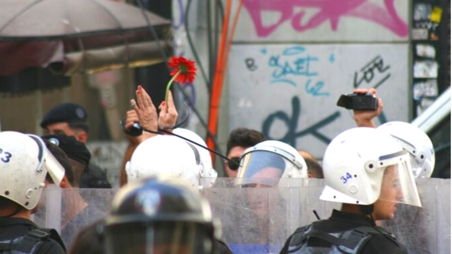 polislerin arasından havaya çiçek kaldıran kelepçeli bir çift el görünüyor
